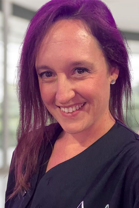 A woman with long, wavy hair dyed purple smiles warmly at the camera, wearing a black jersey with a star design. A blurred indoor background is visible.