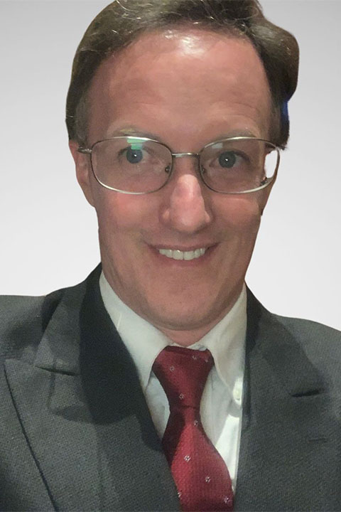 A man with glasses and short hair smiles confidently, wearing a dark suit, white shirt, and a red tie, against a plain light background.