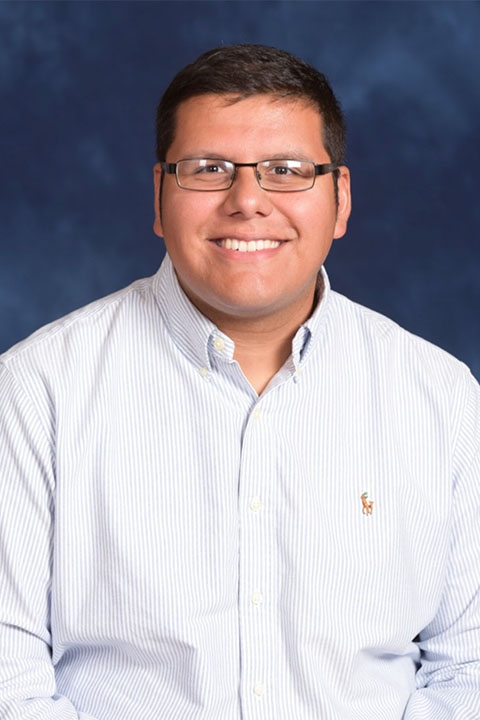 A smiling young man with short dark hair and glasses is seated against a blue background. He is wearing a light blue striped shirt.