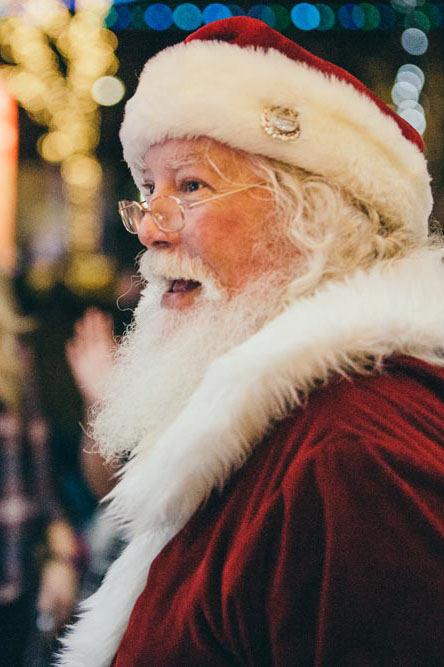 A smiling man with a long white beard and glasses, wearing a traditional red Santa Claus outfit and fur-trimmed hat, stands in a festive, brightly lit environment.