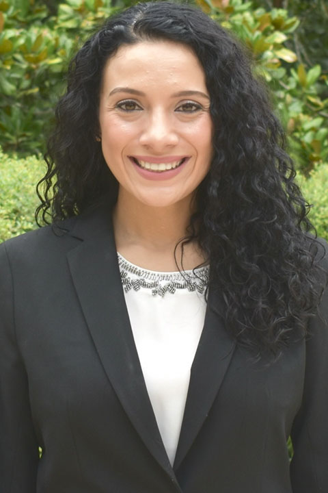 A woman with long, curly black hair is smiling while wearing a black blazer over a white shirt with a decorative neckline. She stands outdoors in front of green foliage.