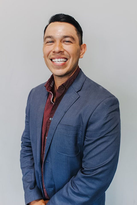 A smiling man in a blue blazer over a maroon shirt stands against a light gray background.