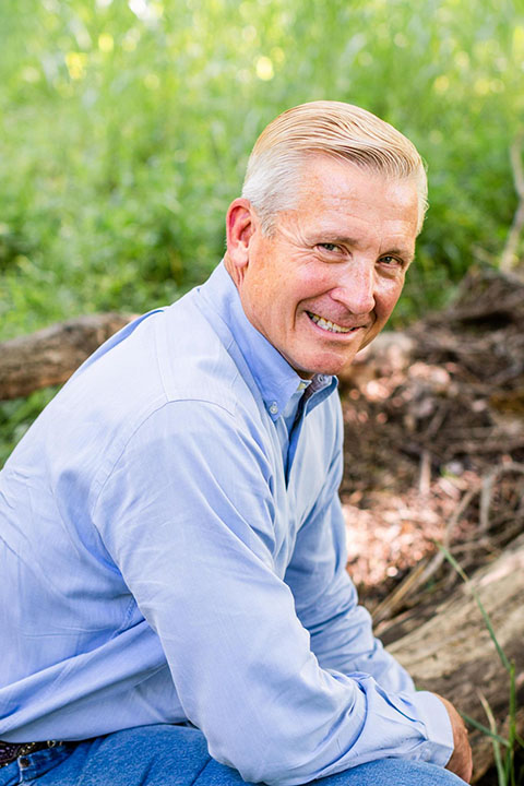 A smiling man with short, light-colored hair wearing a light blue button-up shirt sits outdoors against a backdrop of greenery and fallen logs.