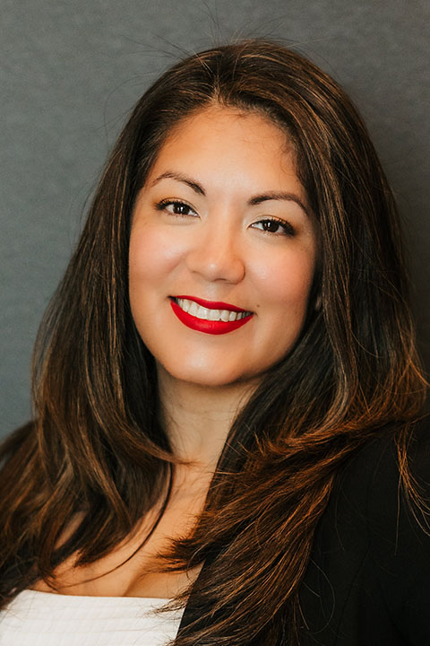 A smiling woman with long, dark hair and bold red lipstick poses against a gray background, wearing a black blazer over a white top.