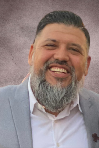A man with a full beard and mustache smiles warmly, wearing a light gray suit over a white shirt. The background features a soft gradient with muted tones.
