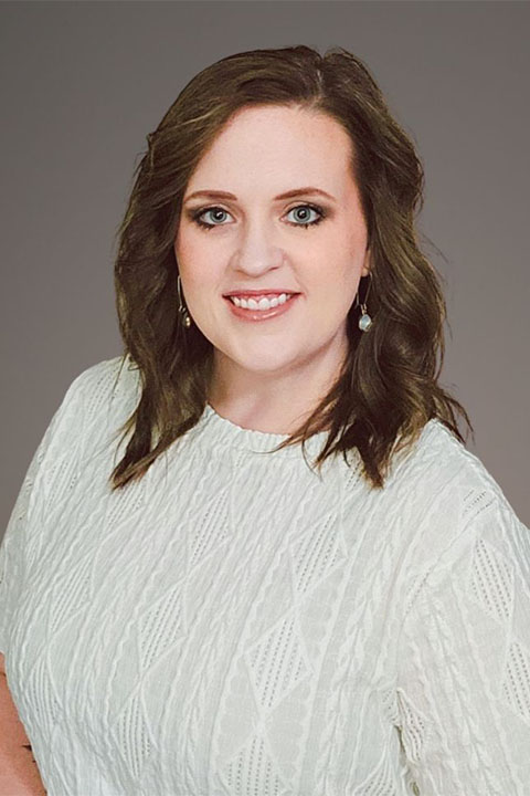 A woman with wavy brown hair and a friendly smile poses in a light patterned top against a gray background. She is wearing earrings and appears confident.