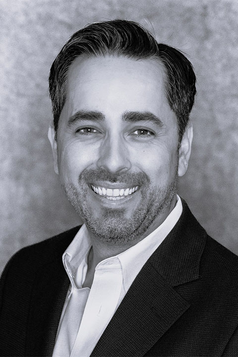 A smiling man with short dark hair and a trimmed beard, wearing a black suit jacket over a white shirt, against a textured gray background.